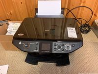 Front view of Epson Stylus RX595 printer showing control buttons, front LCD screen, and paper input tray with paper loaded.