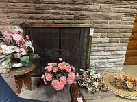 Four faux floral arrangements placed on and in front of a fireplace, small carved wood side table beside them.