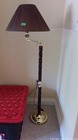Full view of the floor lamp showing the brown pleated shade, dark wood turned post, brass adjustable arm, and base next to a red plastic container.