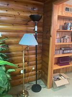 Two floor lamps against a log wood wall: one black torchiere lamp and one brass lamp with a pleated shade and articulating arm, showing overall height and shape.
