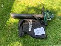 Full view of the green electric Yardworks blower/vac with attached black collection bag and nozzle extension laid out on grass with owner's manual.