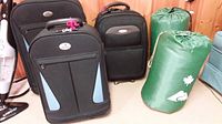 Photo showing three pieces of luggage: one black Aero suitcase and two black and light blue Arkadian suitcases, plus two green sleeping bags in covers.