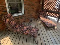 Two loungers with floral cushions and small side table on wooden deck by brick wall.