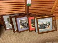 Six framed vintage car prints arranged on the floor, showcasing various colored matting and brown wooden frames.