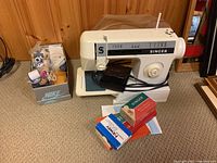 Singer DX-14C sewing machine placed on a beige carpet next to a box containing thread spools, foot pedal in front, and instruction manuals scattered.