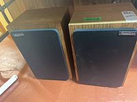 Pair of Electron wood box speakers with black front grille and wood veneer sides placed on floor.