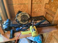 Photo showing Black & Decker circular saw, hand back saw with black handle, Skil electric hedge trimmer, and yellow electric heat tool arranged on a wooden shelf in a workshop.