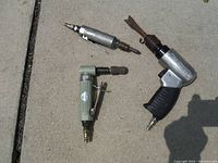 Three pneumatic air tools on concrete surface: two die grinders and one power chisel with attachments.