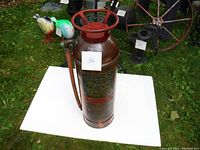 Top and side view of the copper fire extinguisher with red cast iron top and attached copper hose, placed on white board outdoors with garden background.
