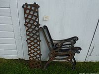 Set of cast iron bench parts including two side arms and two lattice bench slats leaning against white wall on grass
