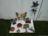 Photo showing all 8 garden decor pieces placed on a white board against an exterior wall with grass in front. Includes butterfly wall art, hummingbird, rooster, sun plaques, palm tree, birdhouse, and teapot.