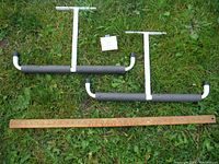 Two ceiling mounted storage racks with white metal frames and foam covered bars laid on grass next to a yardstick for scale.
