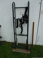 Full view of heavy duty appliance dolly leaning against white shed wall showing black metal frame, two wheels, wooden base and securing straps.