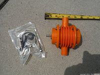 Orange ribbed cylindrical drill pump positioned next to a measuring tape showing approx 5 inch length; small sealed plastic packet of accessories including clamps and wrench visible