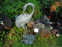Front view of a tall cast iron crane statue standing in a garden near rocks and plants, showing detailed feather texture and weathered paint with some rust spots.