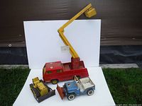 Three vintage metal Tonka toy vehicles: red bucket lift truck, yellow bulldozer, blue jeep with plow, placed on white background