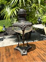 Front view showing full lamp with detailed metal base and cut glass body, placed on wooden surface outdoors with foliage background.