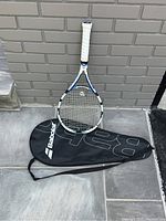 Babolat Drive Lite tennis racquet standing against a brick wall with black Babolat cover lying horizontally in front of it.