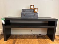 Black modern console table against a beige wall and wooden floor, showing top surface with decorative boxes and candles (not included) and lower shelf.