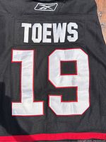 Back view of black hockey jersey showing 'TOEWS' and number 19 in white with red border, made by Reebok.