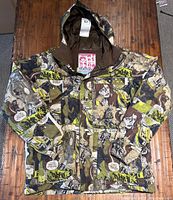 Front view of Burton snowboard jacket laid flat, showing hood, full zipper, chest pocket, and comic-style camouflage pattern.