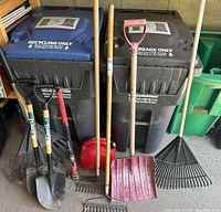 Photo showing collection of gardening hand tools against recycling and garbage bins, including leaf rakes, shovels, small rake, and a red red Jerry can