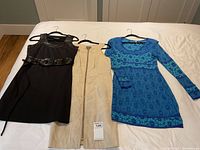 Three ladies dresses laid flat on a bed, showing black sleeveless dress with belt, beige zip-up dress, and blue floral long-sleeve dress.