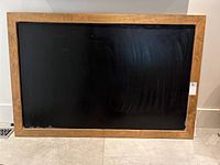 Frontal view of large framed blackboard with oak frame standing against a light-colored wall on a tiled floor. Black surface shows some marks.