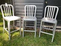 Full view of three modern aluminum bar stools arranged side by side outdoors on grass against a dark siding wall showing design and condition.
