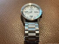 Men's Nixon stainless steel watch face showing white dial with black numeric markers and silver hands.