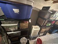 View of two black plastic shelving units in a basement or garage setting holding various storage bins and miscellaneous items not included in sale.