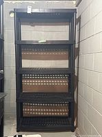 Front view of the entire black plastic shelving unit standing against cinder block wall on concrete floor.