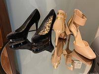 Photo showing two pairs of women's high heel shoes on a glass table reflecting their image. Black velvet Stuart Weitzman shoes on the left, nude Nine West shoes with gold accents on the right.