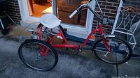 Full side view of adult red tricycle with three wheels, white cushioned seat, and part of rear basket visible.