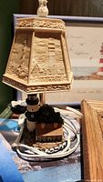 Close-up photo of a lighthouse-themed desk lamp showing beige embossed lampshade with lighthouse and building designs, decorative finial on top, and miniature lighthouse with house and landscaping on base.