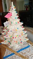 Full view of the 23-inch white ceramic Christmas tree with multicolor removable bulbs on each branch tip, sitting on a lace-covered table with a measuring stick showing height.