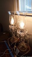 Two crystal table lamps sitting on a surface near a window, both lamps lit, showing the design and crystal drops.