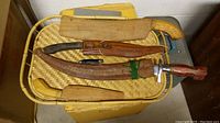 Photo showing four Filipino cutting utensils with their sheaths laid on a woven tray. One black-handle pocket cutter among three machete-style knives with wooden and leather sheaths.