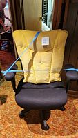 Front view of black desk chair with large tufted yellow cushion attached to backrest, showing chair seat, armrests, and base.
