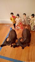 Photo showing four Filipino dolls standing upright behind two wooden water buffalo figurines on a wooden floor, highlighting the dolls' traditional attire and the buffaloes' carved details.