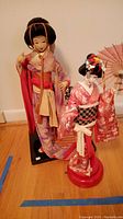 Both oriental dolls shown together on wooden floor, one taller doll in purple kimono and shorter one in red kimono on musical stand holding a parasol.