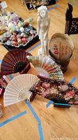 Group of five decorative handheld personal fans with holders in various colors and floral patterns placed on floor near a basket.
