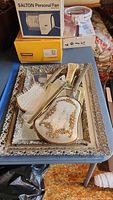 Vintage dresser set including hand mirror, comb, hairbrush, clothes brush on mirror tray, Salton personal fan box, round vintage hair dryer, and heat wrap.