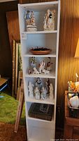 White tall bookcase with several porcelain figurines arranged on shelves, including a pair figurine on the top shelf and miniature military figures below.