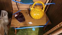 Photo of a small wooden table holding a red stained glass mask and a yellow ceramic pitcher.