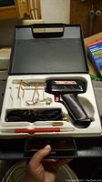 Electric soldering gun with accessories inside molded plastic case with handle, black tool with power cord and metal stands.