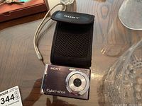Sony Cyber-shot digital camera next to protective carrying case on wood surface.