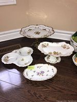 All four ceramic pieces shown on a hardwood floor including two pedestal plates and two serving trays with floral and gold detailing.