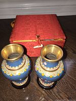 Top and front view of two small blue cloisonne vases showing floral and cloud motif decoration, with brass rims, positioned on a wooden floor next to a red decorative storage box.