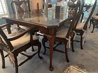 Photo showing the corner of the wooden dining table with claw foot legs and the custom glass top cover, plus some of the matching dining chairs with patterned cushions.